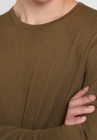 Braunes Langarm-Strickshirt mit Rundhalsausschnitt, das einen strukturierten Stoff und gerippte Akzente am Hals und an den Schultern aufweist.