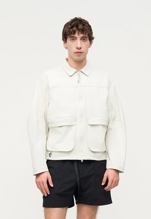 Giovane uomo che indossa una giacca bianca con zip e grandi tasche frontali e pantaloncini neri su uno sfondo chiaro uniforme.