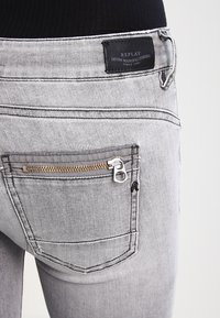 Los jeans de mezclilla gris tienen un bolsillo con cremallera en el lado derecho. Una etiqueta negra muestra el nombre de la marca "REPLAY". Las costuras son de color gris claro.