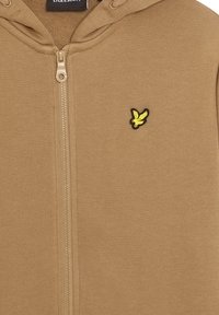Khaki zip-up hoodie gjord av mjukt tyg, med en broderad logotyp i gult och svart på bröstet och huva med dragsko.