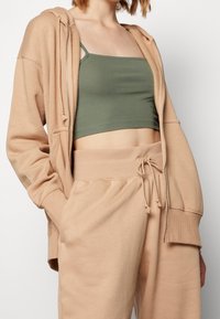 Person som bär en beige zip-up hoodie och matchande byxor med dragsko, i kombination med en olivgrön kortärmad topp.