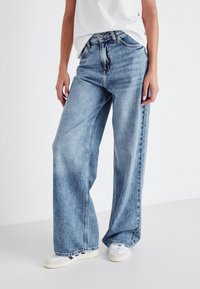Personne portant un jean bleu clair à jambes larges, des baskets blanches et un haut blanc ample, debout devant un fond uni.
