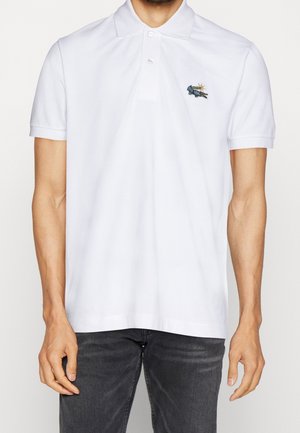 Mies, joka pukeutunut valkoiseen lyhythihaiseen polopaitaan, jossa on kaulus, kaksi nappia ja vasemman rinnan pienessä brodeerattu krokotiililogo.