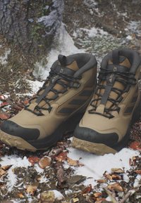 Braune und schwarze Wanderschuhe mit schwarzen Schnürsenkeln auf verschneitem, blattbedecktem Waldboden nahe einem Baumstamm.