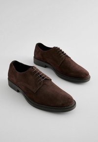 Zapatos de ante marrón con cordones, puntera redonda y una suela de goma negra en contraste. Presentan detalles de costura a lo largo de la suela y la lengüeta.