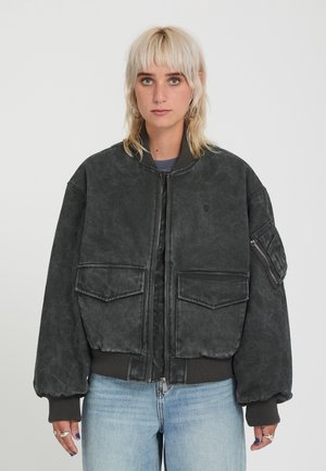 Persona bionda con anello al setto nasale che indossa una giacca bomber nera oversize con grandi tasche frontali e jeans azzurri chiari, in piedi su sfondo semplice.