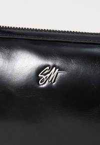Juodas odinis maišelis su lygiu paviršiumi, turintis sidabro "SM" logotipą. Uždarymas užtrauktuku viršuje su metaliniais akcentais.