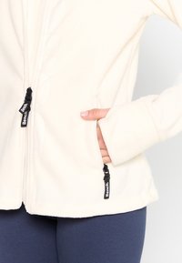 Personne portant une veste polaire crème avec des tirettes de fermeture éclair noires de la marque Bench, main dans la poche zippée droite, portée avec un pantalon bleu foncé.