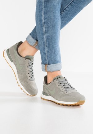 Osoba ubrana w podwinięte niebieskie dżinsy i szare buty Nike stojąca na białej podłodze.