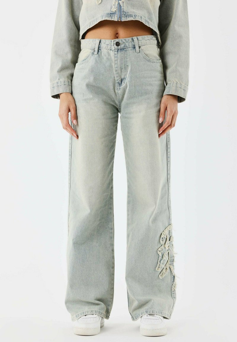 Jean en denim bleu clair à jambes larges présentant des accents usés sur les côtés et un design traditionnel à cinq poches. Présenté avec des baskets blanches.