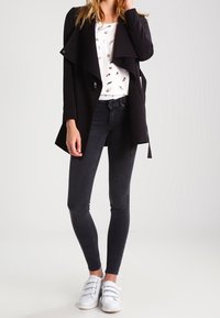 Un abrigo trinchera negro de manga larga, superpuesto a una blusa blanca con un pequeño estampado, combinado con unos jeans ajustados oscuros y zapatillas blancas tipo slip-on.