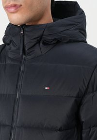 Black puffer jacket with a hood, featuring stitched panels, a front zipper, and a small logo in red, white, and blue on the chest.