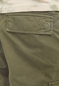 Detailní záběr na zelené látkové cargo kalhoty s obdélníkovou zadní kapsou se klopou a viditelným šitím.