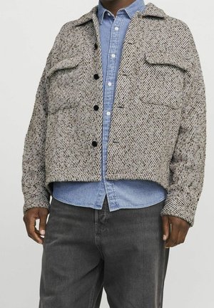 Mand iført en struktureret beige jakke med knapper over en lyseblå denimskjorte og mørkegrå bukser, stående mod en ensfarvet baggrund.