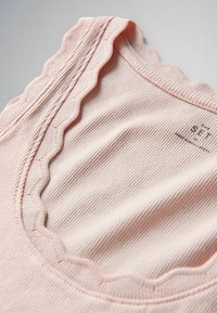 Detailný záber na bledo ružový pruhovaný tkaninový výstrih s vlnitým lemovaním a malým potlačeným štítkom.