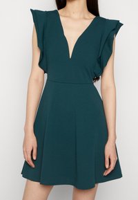 Tyrkysové šaty s hlubokým výstřihem do V, volány na ramenou, přiléhavý pas a rozšířená sukně. Vyrobeno z hladkého, elastického materiálu.