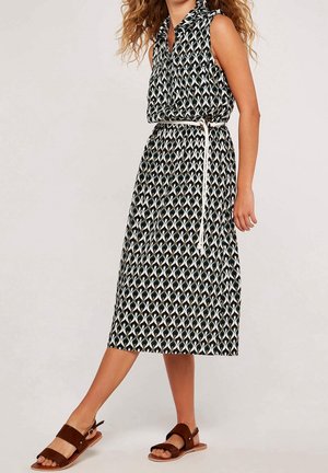 Femme portant une robe midi sans manches à motifs avec une ceinture blanche et des sandales marron, debout avec une jambe légèrement levée.
