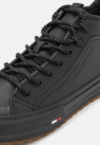 Svart lærersneaker med gummisåle, har teksturerte detaljer, flate sorte snørebånd og et tri-farget logo detaljer nær sålen.