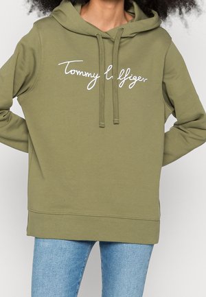 Osoba ubrana w oliwkowo-zieloną bluzę z kapturem z białym, ozdobnym napisem "Tommy Hilfiger" oraz jasnoniebieskie dżinsy, trzymająca ręce za plecami.