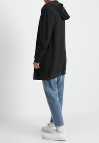 Person in einem langen schwarzen Kapuzencardigan, hellblauen, verkürzten Jeans, weißen Socken und weißen Turnschuhen, stehend vor einem schlichten Hintergrund.