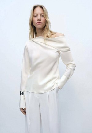 Blusa crema a spalle scoperte con maniche lunghe, caratterizzata da un accento nero ai polsini, realizzata in un tessuto liscio e lucido; abbinata a pantaloni bianchi.