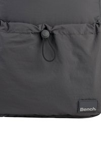 Gray nylon backpack with a drawstring closure, featuring a black rubberized logo patch and smooth texture.