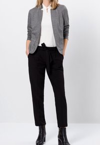 Veste à motifs gris sur un haut blanc, associée à un pantalon taille haute noir et des bottines noires à chevilles. Comprend des poches et une taille nouée.