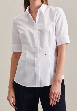 Mujer que lleva una camisa blanca de manga corta con botones y un pequeño bordado de rosa negra en la parte delantera, combinada con pantalones oscuros.
