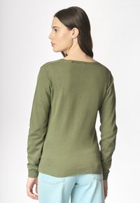 Una donna con capelli di lunghezza media indossa un maglione verde oliva e pantaloni azzurri chiari, in piedi con la schiena rivolta verso l'osservatore su uno sfondo neutro.