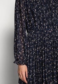 Laivastonsininen mekko, jossa on kermaisen värisiä kukkakuosisia, pitkät hihat, joustava vyötärö, nappikiinnitys ja valuva hame.