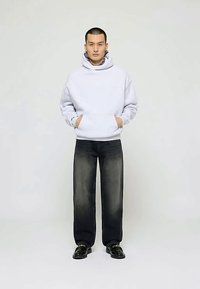 Muškarac stoji s rukama u džepovima, odjeven u svijetlosivu duksericu, izblijedjele crne traperice i crne kožne mokasine na jednostavnoj pozadini.