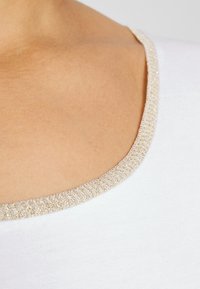 Hvid skjorte med rund halsudskæring og en tekstureret gylden kant. Stoffet ser glat ud med en let glans.