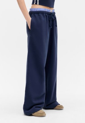 Navy sweatpants with a loose fit, elastic waistband featuring a blue checkered interior, and side pockets. Textured fabric with soft drape.