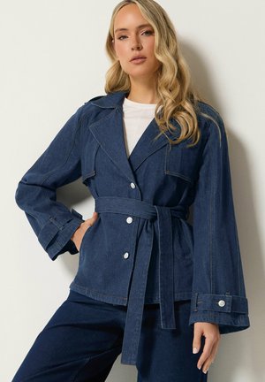 Kvinde med langt bølget blondt hår iført en mørk denimjakke med bælte og sølvknapper samt matchende bukser, poserer mod en lys væg.