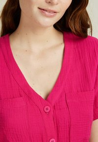 Chemise rose à boutons avec un col en V, en tissu texturé et dotée de deux poches. Elle a des boutons plats et une coupe décontractée autour de l'encolure.