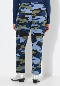 Cargo-Hosen mit einem blau, schwarz und grün gemusterten Camouflage-Design. Sie bieten eine lockere Passform, seitliche Taschen und eine glatte Stofftextur. Silberne Stiefeletten.