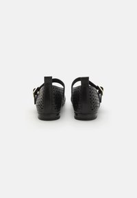 Sandalias de cuero negro con un diseño perforado, cierre con hebilla ajustable y un tacón bajo. La vista trasera muestra el detalle de la correa en el tobillo.