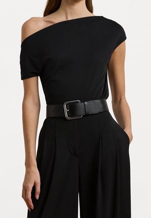 Vrouw draagt een zwarte one-shoulder top, ingestopt in zwarte, hoog getailleerde broek met wijde pijpen, met een brede zwarte riem met zilveren gesp.