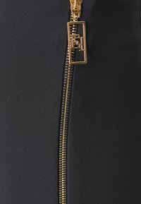 Robe noire en tissu lisse, mise en valeur par une fermeture éclair dorée avec une languette rectangulaire et un logo le long de la couture.