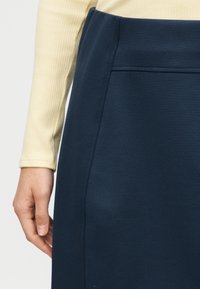 Χέρι που ακουμπά δίπλα σε σκούρο μπλε φούστα με διακριτική λεπτομέρεια ραφής, φορεμένη με μπεζ μακρυμάνικη μπλούζα με ριμπ ύφασμα.