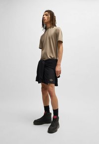 Chemise beige à manches courtes, shorts noirs avec détails à motifs, baskets montantes noires et chaussettes noires. Tissu lisse, coupe décontractée.