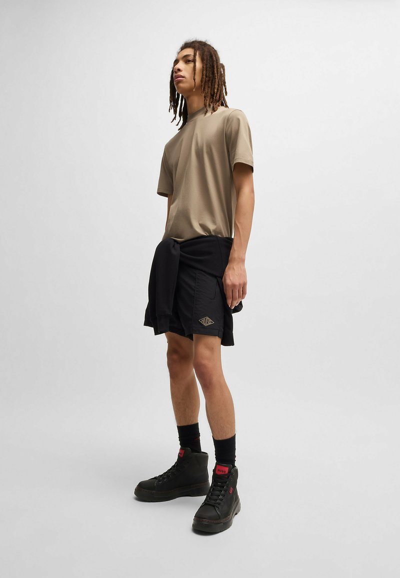 Chemise beige à manches courtes, shorts noirs avec détails à motifs, baskets montantes noires et chaussettes noires. Tissu lisse, coupe décontractée.