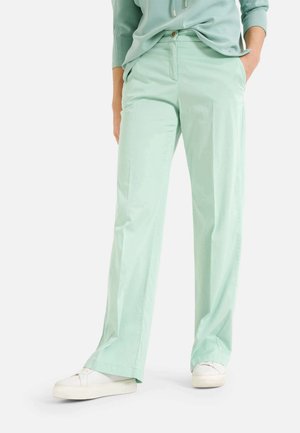 Person iført lysegrønne bukser med lige ben, med hænderne i lommerne, kombineret med hvide sneakers og en matchende lysegrøn top.