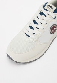 Sneaker bianco con materiali in mesh testurizzato e suede, dettagli blu navy e una patch rotonda con logo. Presenta un laccio prominente e una suola in gomma.