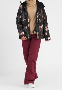 Blommönstrad svart skidjacka med huva, bärs över en beige polotröja, kombineras med vinröda snöbyxor. Vita skor fulländar outfiten.