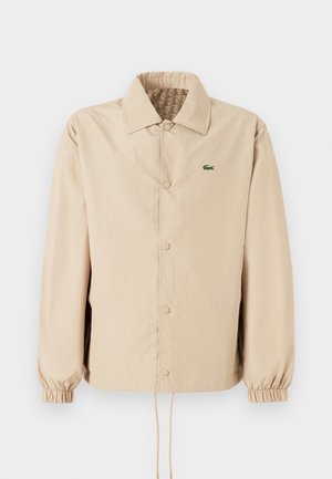 Beige lehká bunda s límečkem, zapínáním na knoflíky, elastickými manžetami a stahovacími okraji. Na levé straně hrudi má zelené logo.
