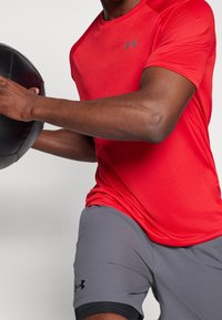 Červené sportovní triko s kulatým výstřihem a přiléhavým střihem, spárované s šedými šortkami s černým akcentem. Drží černý cvičební míč.