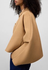 Kameelwollen jas met een oversized silhouet, voorzien van een capuchon, knoopdetails en een zachte textuur. Eenvoudig ontwerp zonder zichtbare patronen.