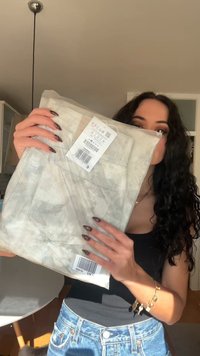 Bež vezene kavbojke z barvnim vzorcem v prozorni embalaži, z zadnjim žepom in etiketo z črtno kodo. Kavbojke so vidne od spredaj.