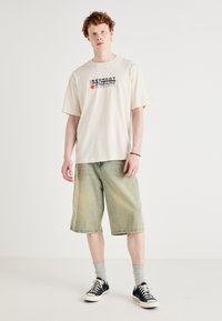 Creme farvet bomulds t-shirt med sort og rød grafisk tekst, kombineret med lysegrønne denim shorts. Sorte sneakers og grå sokker fuldender looket.